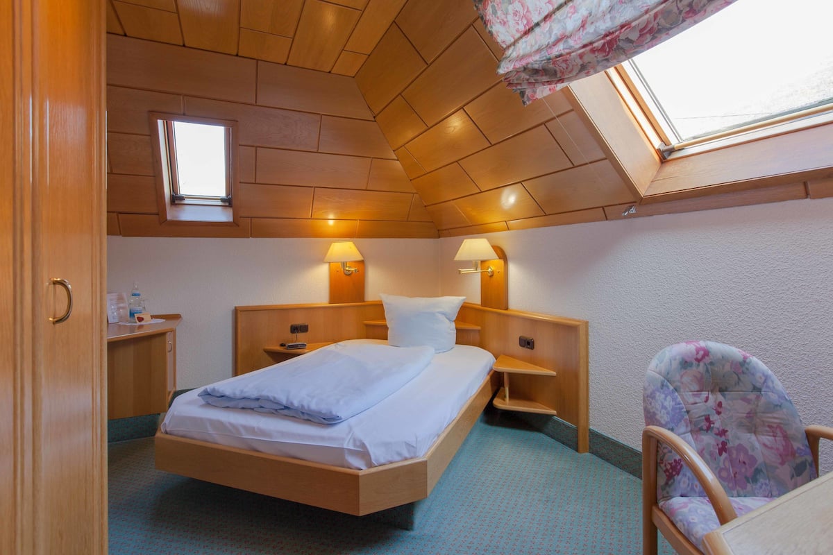 A small, cozy room with a wooden ceiling, a single bed, two bedside tables with lamps, a chair, and a window with a view.