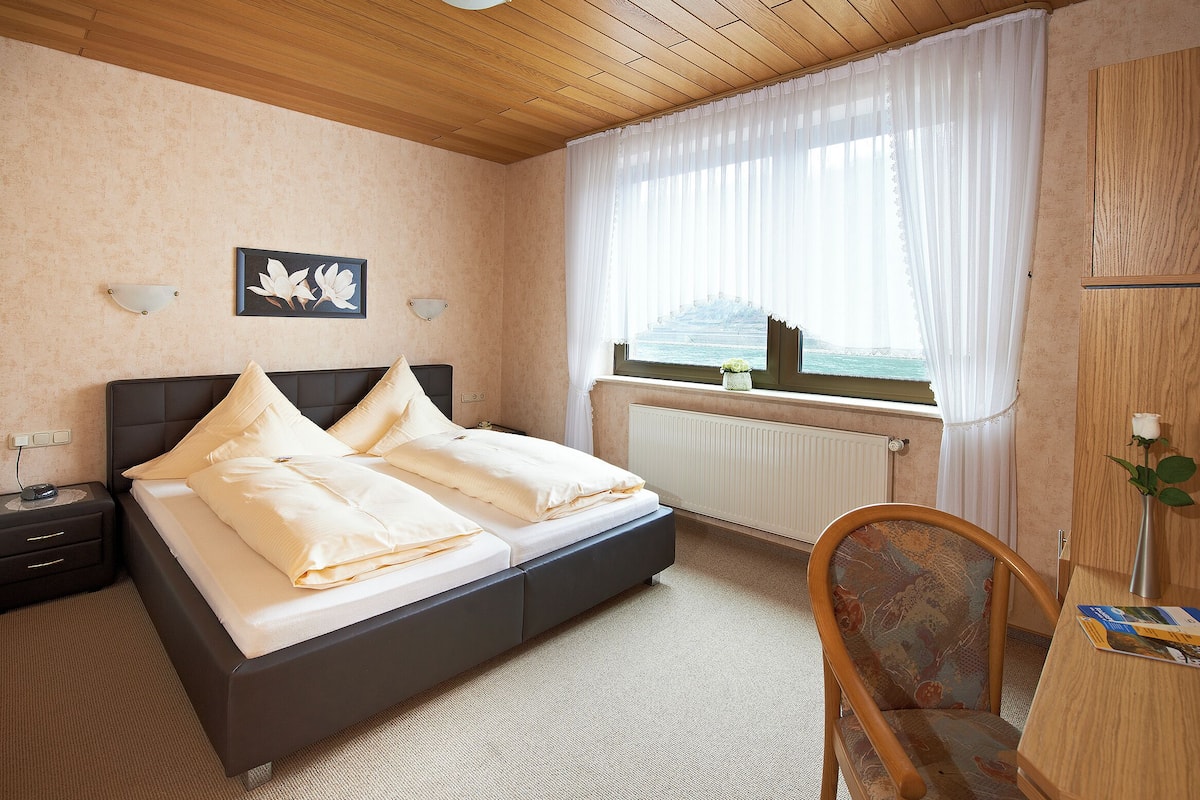 A bedroom with a bed, a nightstand, a chair, a window with curtains, and a view of the sea.
