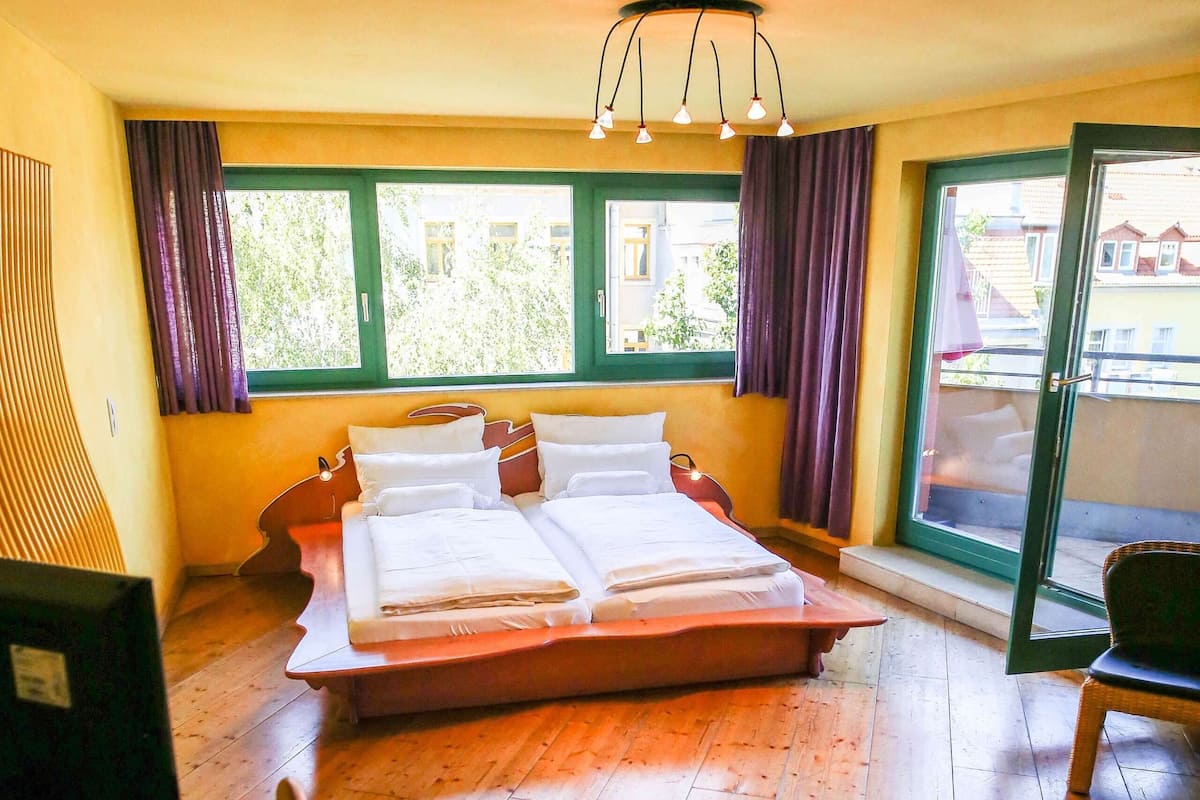 A hotel room with a large bed, a desk, and a chair. There are windows with curtains, a ceiling light, and a view of the outside.