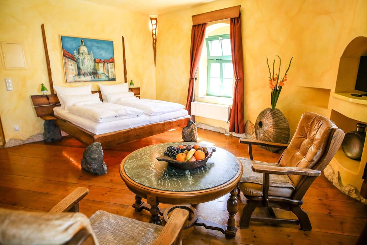 A room with a bed, a round wooden table with a bowl of fruit, a leather chair, and a painting on the wall.