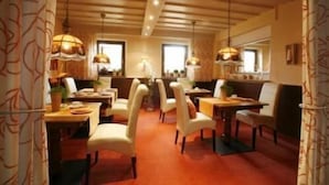A dining area with wooden tables and chairs, pendant lighting, and a view of the kitchen in the background. - Hotel Zur Linde (Hanau)