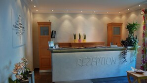 A reception desk with a 'RECEPTION' sign, potted plants, a wooden cabinet, and a wall with a welcome sign. - Hotel Zur Linde (Hanau)