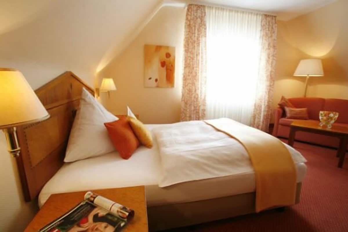 A hotel room with a bed, a nightstand, a lamp, a sofa, and a window with curtains.
