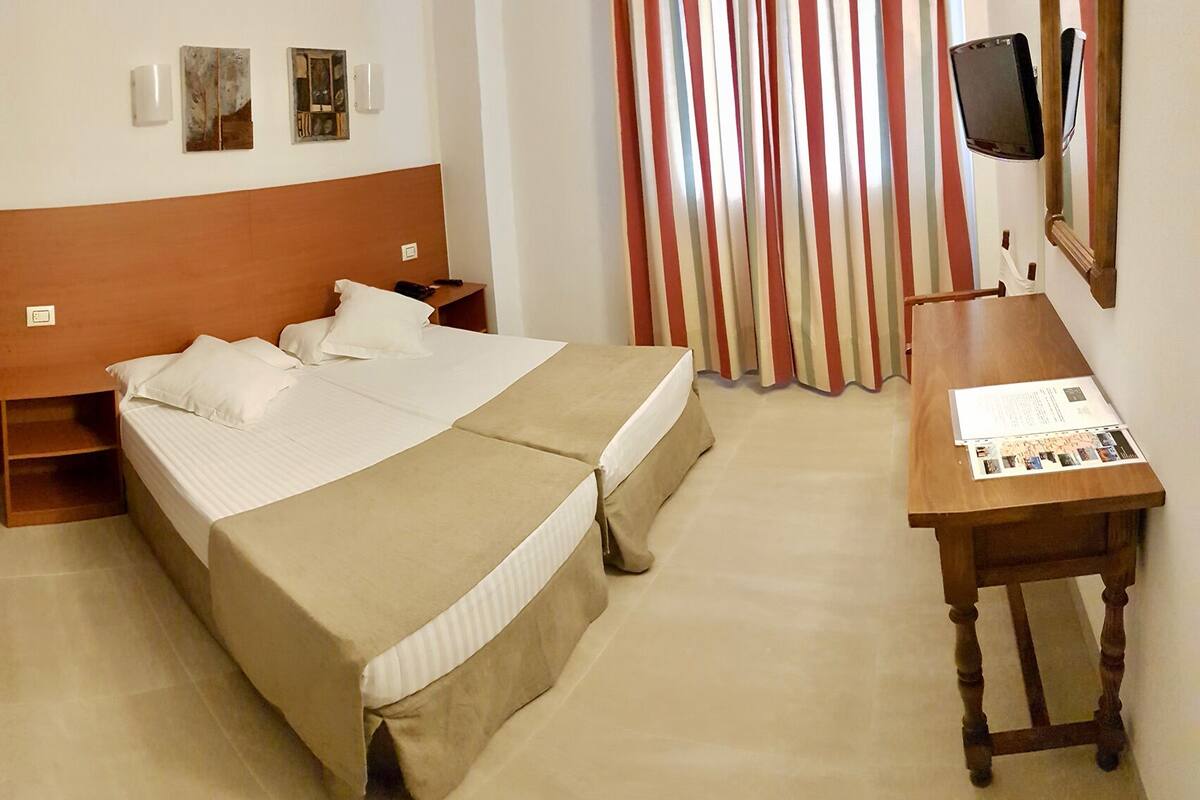 Habitación de hotel con dos camas, cabecera de madera, escritorio con silla, televisor y ventana con cortinas a rayas.