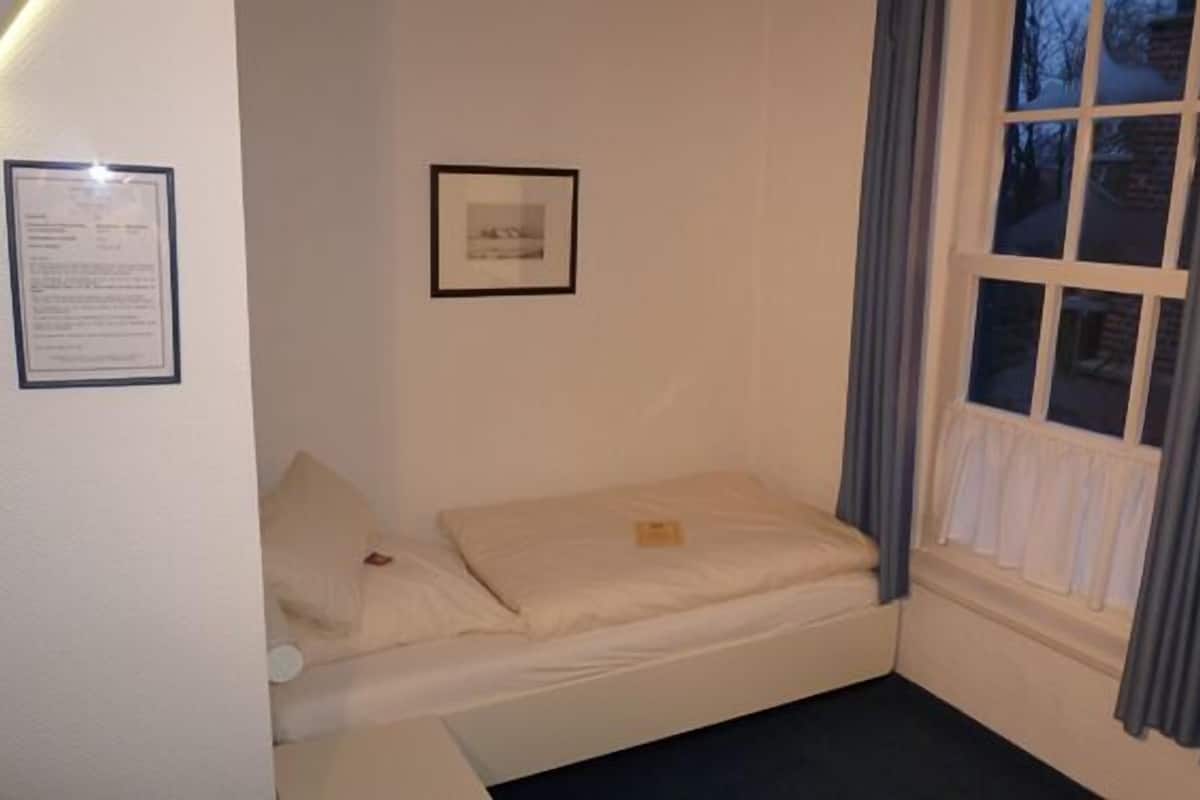 Une petite chambre avec un lit simple, une fenêtre, un tableau encadré au mur et un tableau d’affichage.