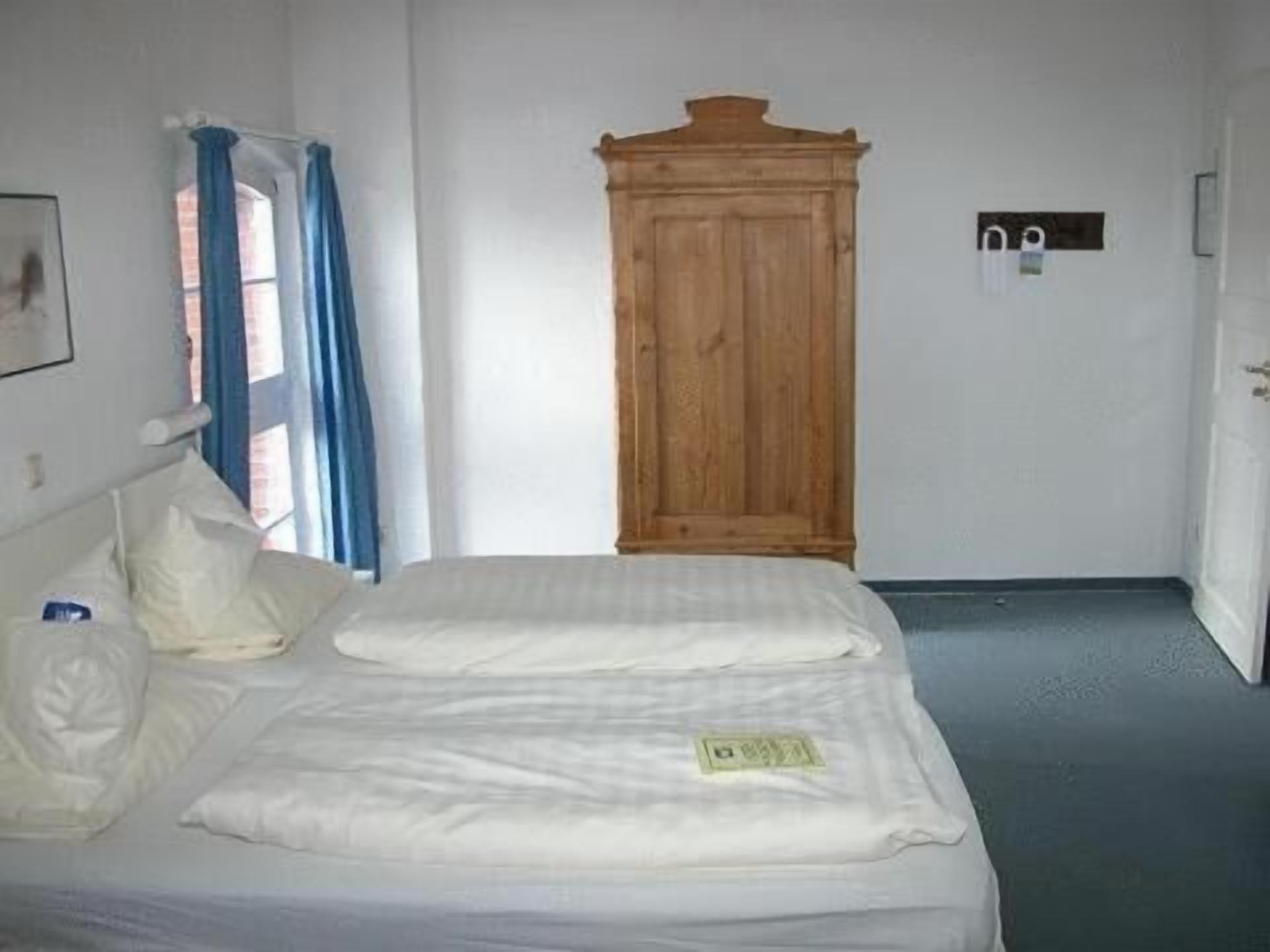 A single-bed room with a wooden wardrobe, a window with blue curtains, and a bedside table with a lamp.