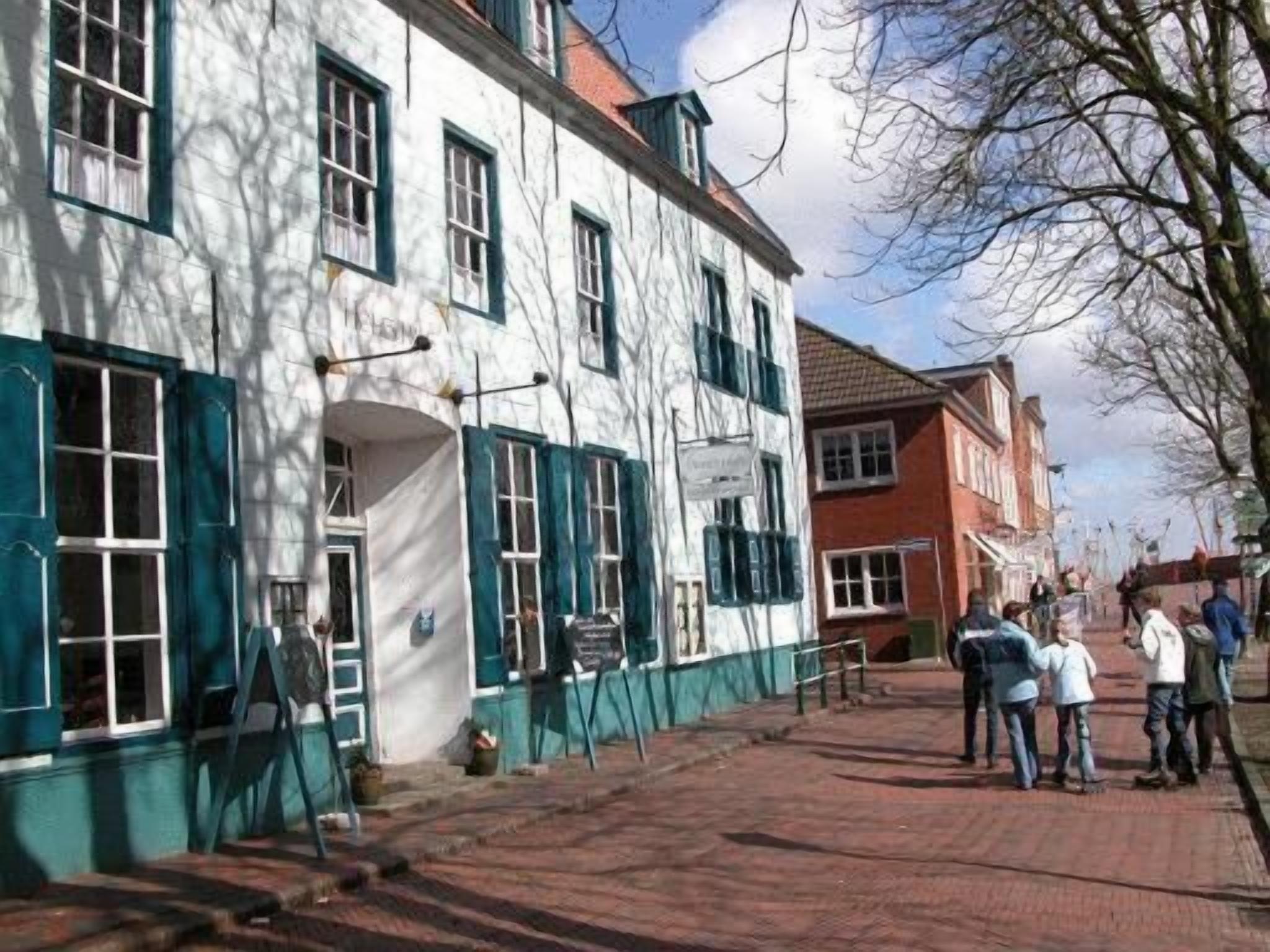 Un edificio con fachada blanca y contraventanas verdes, un camino de ladrillo y personas de pie en la acera.