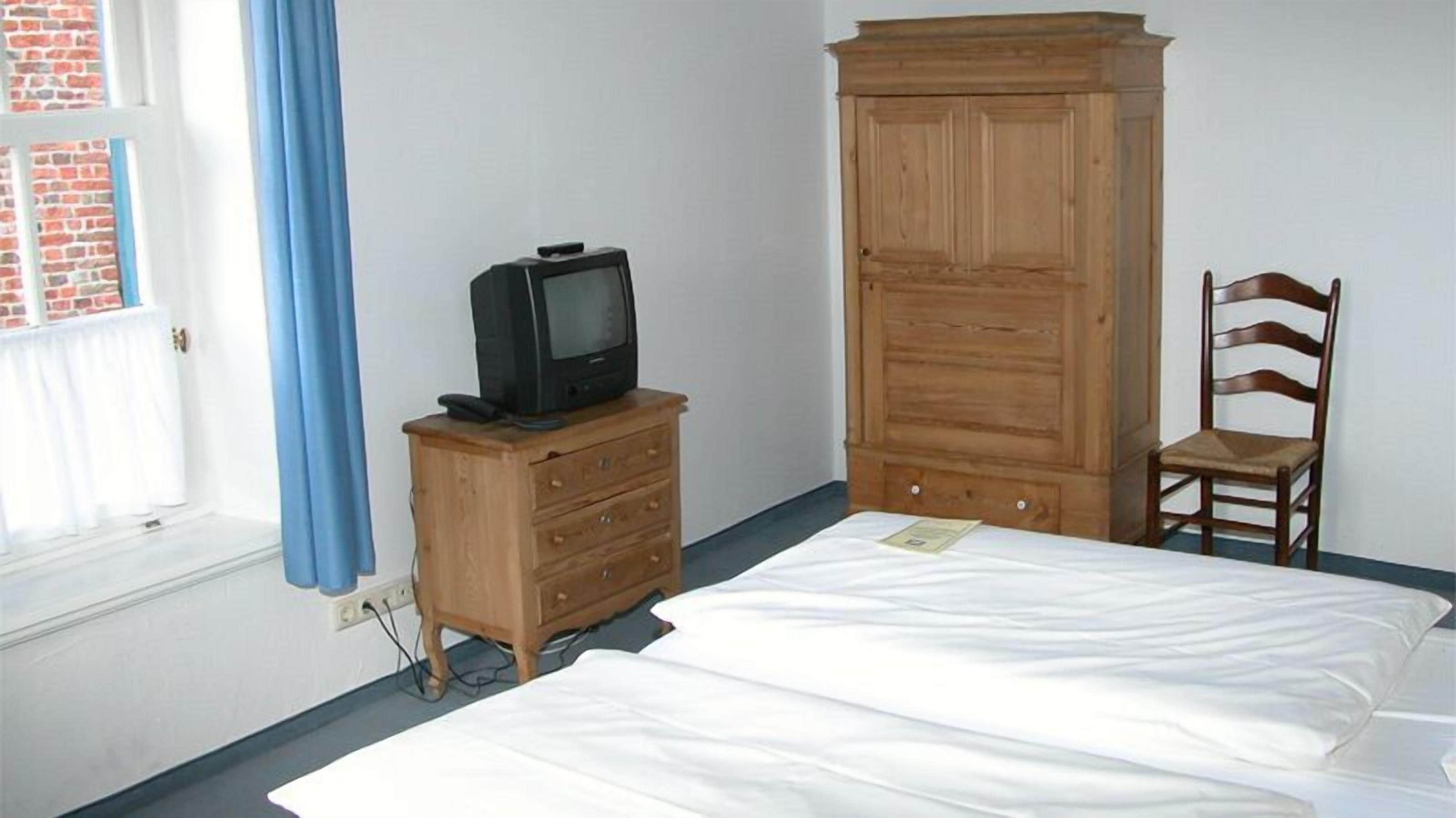 Un dormitorio con una cama, un aparador de madera con televisor, un armario de madera, una silla de madera y una ventana con cortinas azules.