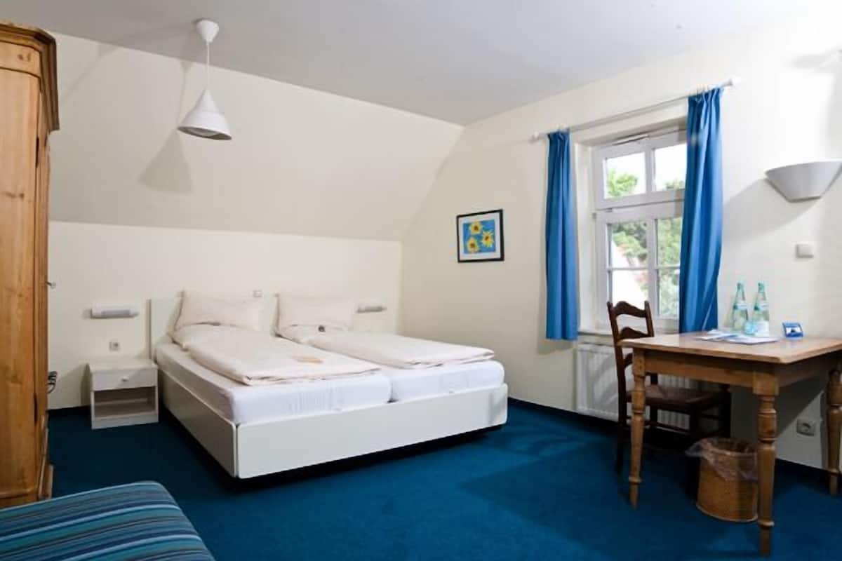 Une chambre à coucher comprenant un lit, un bureau, une chaise, une fenêtre avec des rideaux, un tableau au mur et un luminaire suspendu.