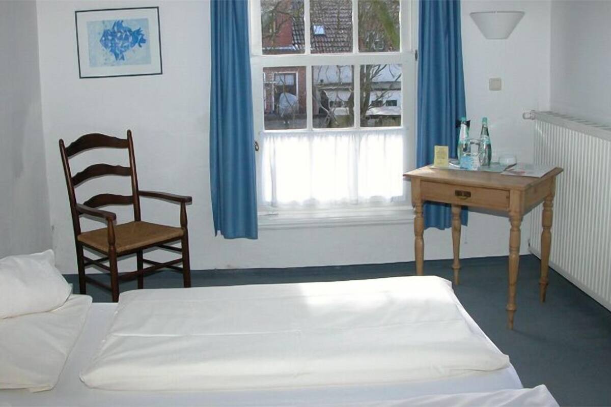 Une chambre à coucher avec un lit, une chaise en bois, une petite table avec une lampe et une fenêtre avec des rideaux.