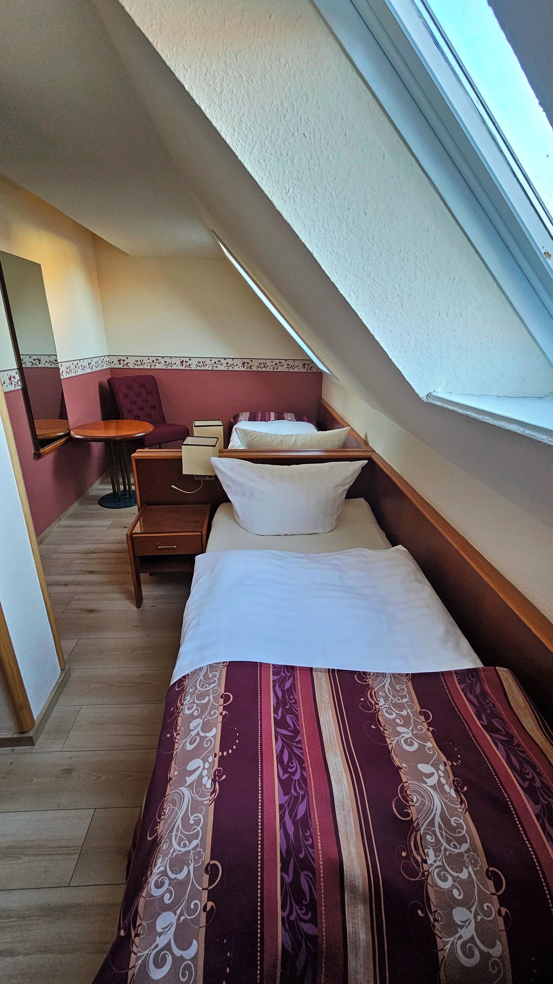 A small, cozy room with a sloped ceiling, a bed with white linens, a wooden nightstand, a small table, and a chair.
