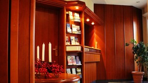 Een receptiegedeelte met een houten receptiebalie, een boekenkast met tijdschriften en een potplant.