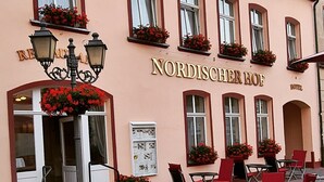 Een gebouw met het bord "NORDISCHEER HOF", wat een hotel aangeeft. Er staan buitentafels en stoelen, en bloembakken op de ramen.