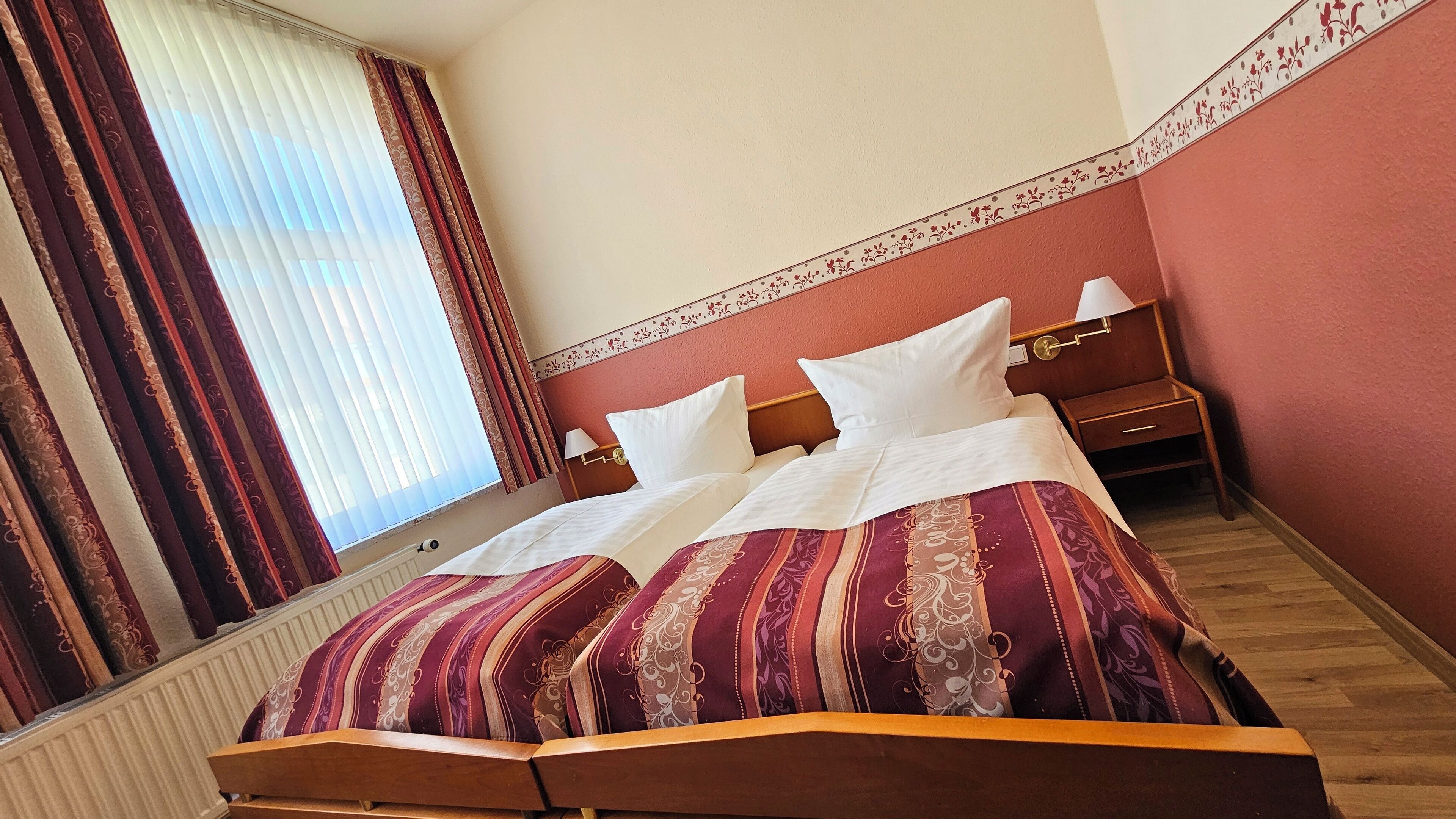 A hotel room with a bed, curtains, a nightstand, and a wall with a floral border.
