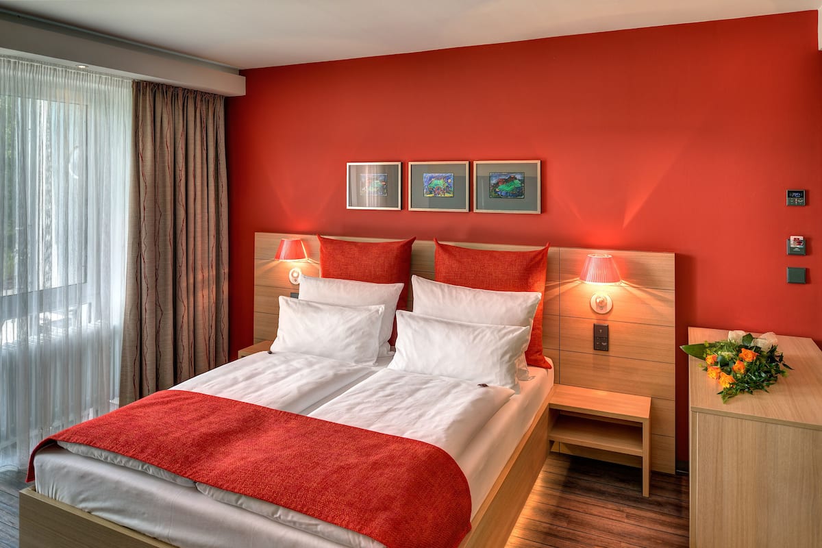 Ein Doppelbett mit weißer und roter Bettwäsche, zwei Nachttische mit Lampen sowie ein Holznachttisch mit Blumen in einer Vase.