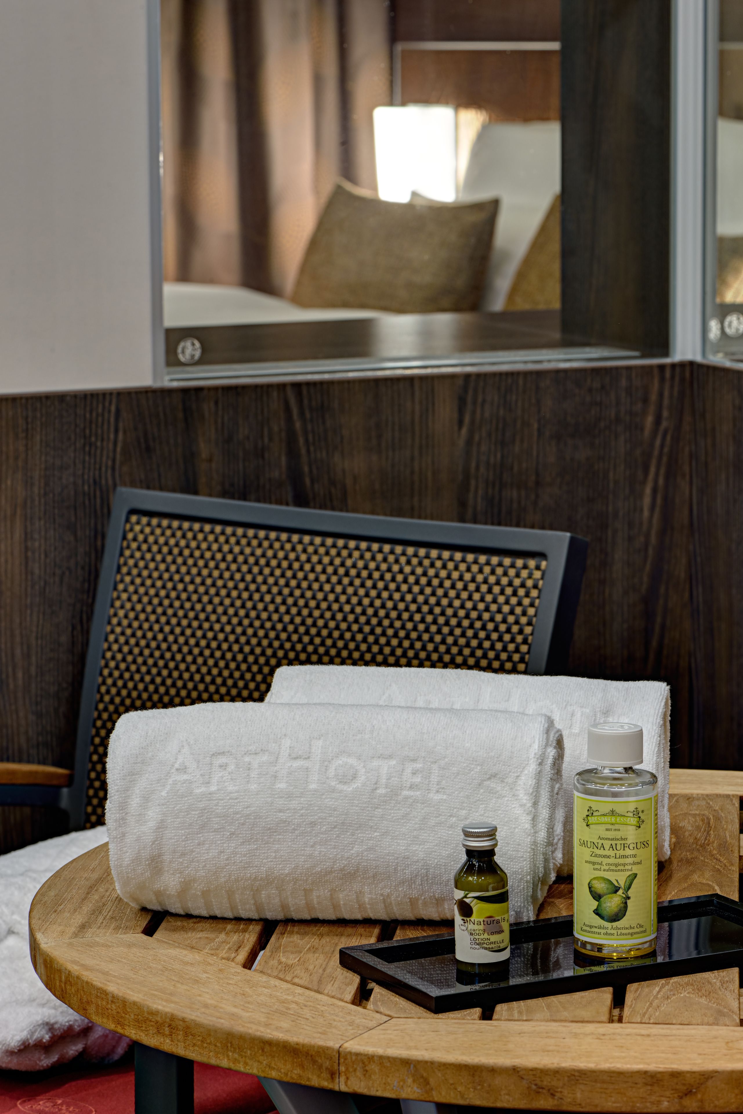 A hotel room with a bed, a wooden headboard, a round wooden table with a towel, a bottle of natural oil, and a bottle of sauna oil.