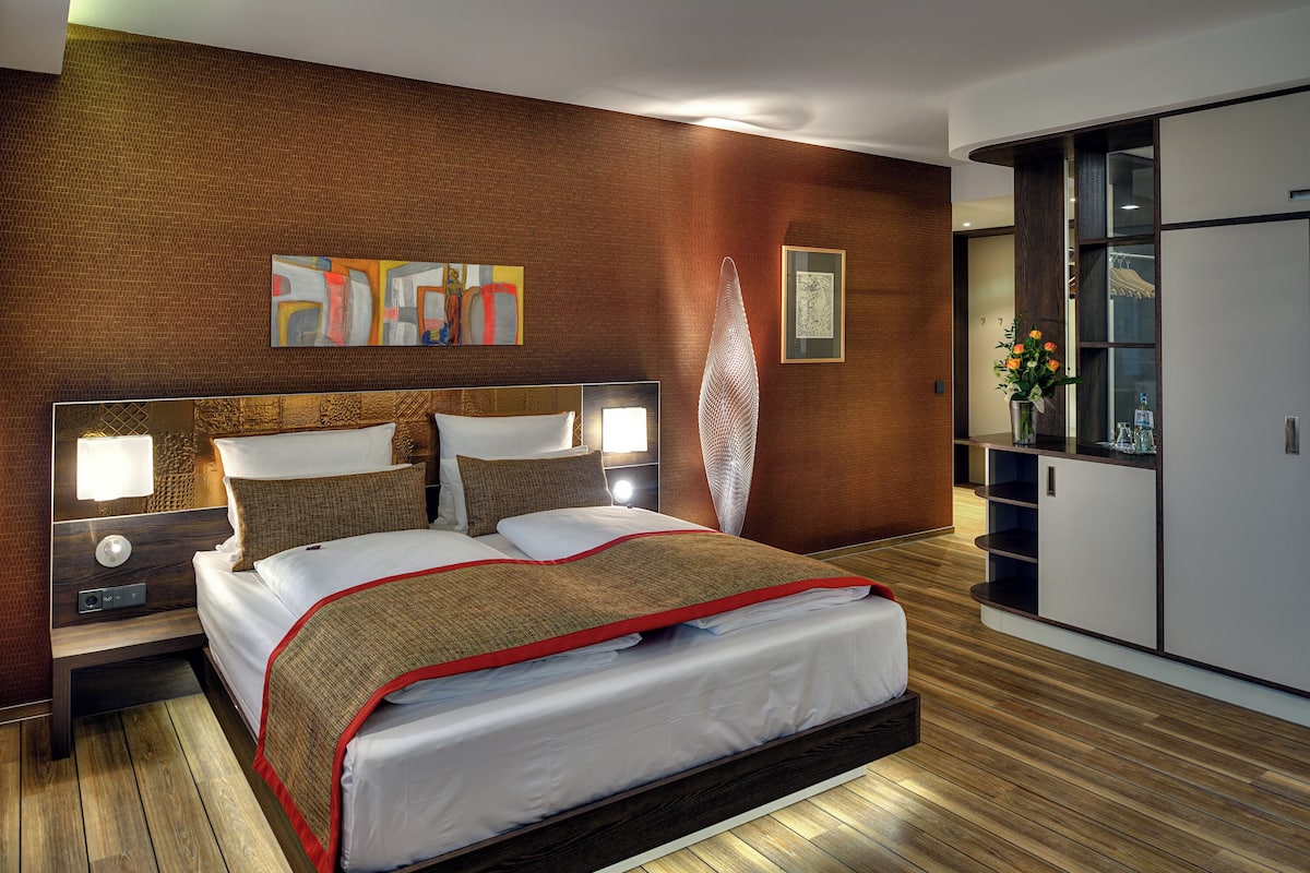 Ein modernes Hotelzimmer mit einem großen Bett, Holzboden, einem Nachttisch mit Lampe, einem Kleiderschrank und Bildern an der Wand.