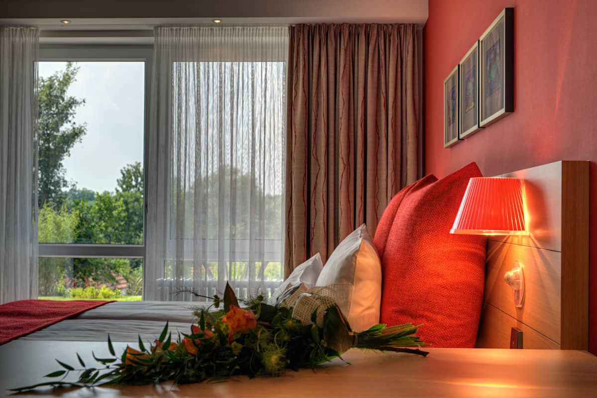 Ein Hotelzimmer mit einem großen Bett, einer Nachttischlampe, einem Fenster mit Vorhängen und Blick auf Grün.