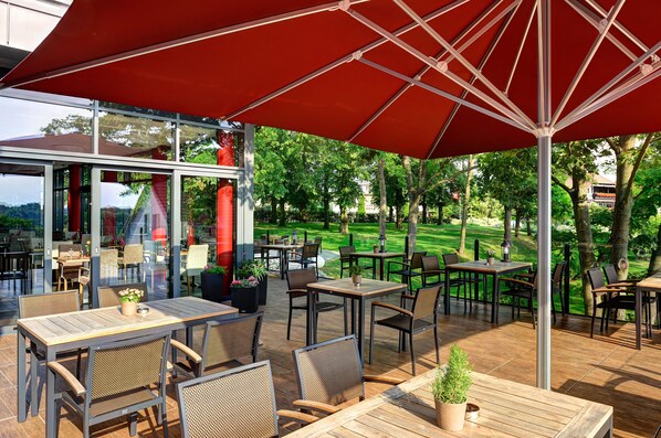 Un patio exterior con mesas y sillas bajo un gran paraguas rojo, con vistas a un césped y árboles bien cuidados.