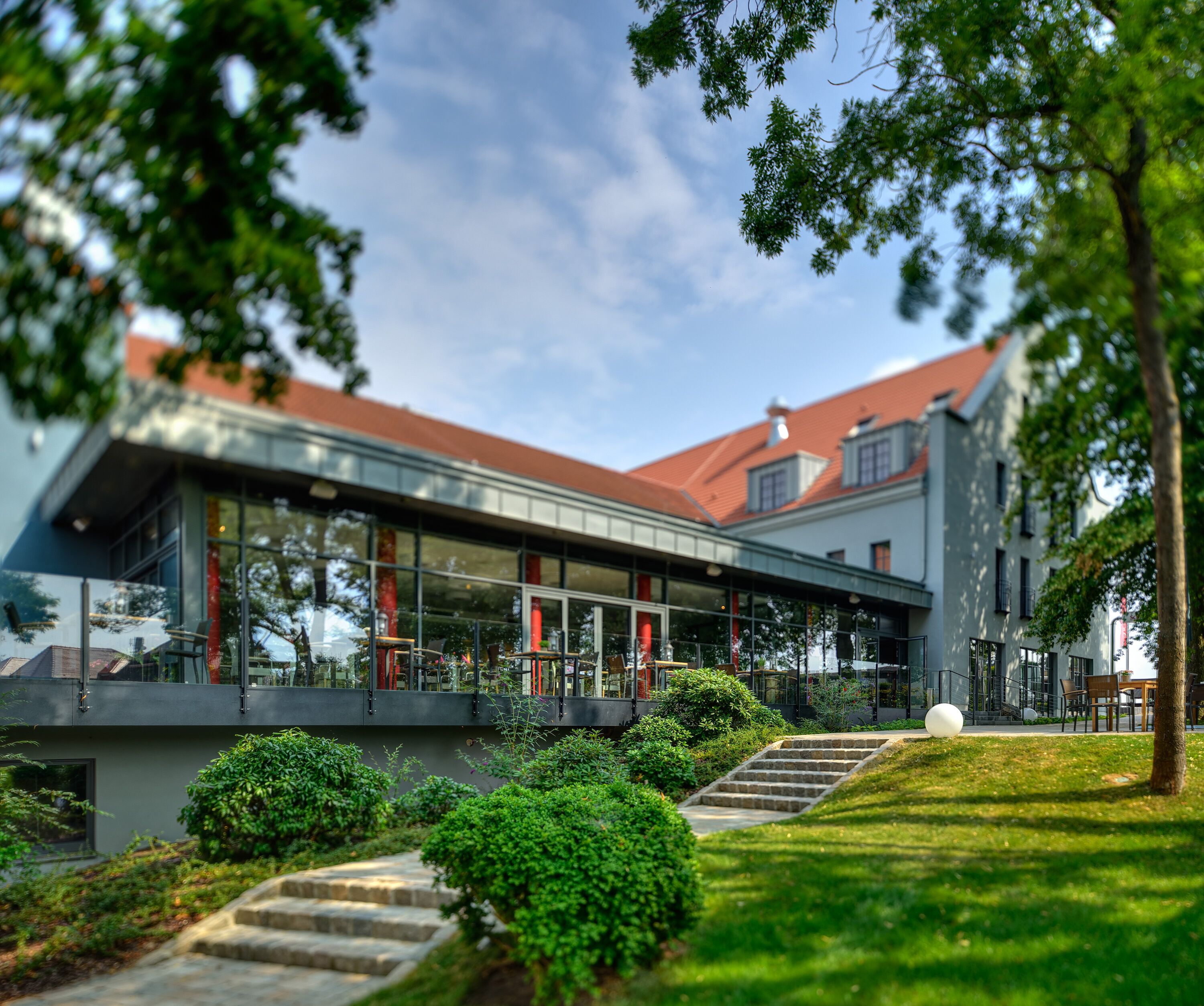 A modern building with a glass facade, a red roof, and a well-maintained garden with a pathway leading to the entrance.
