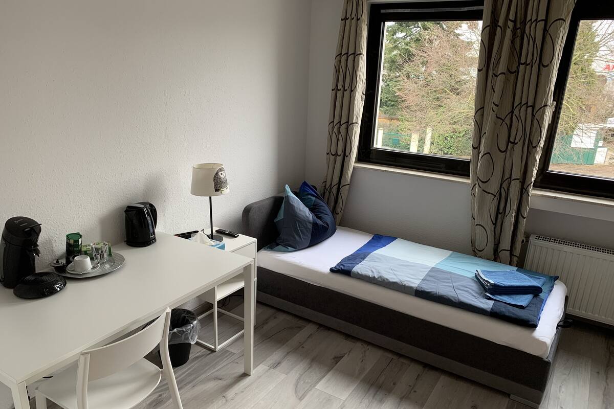A small, tidy room with a single bed, a desk, a chair, a lamp, and a window with curtains.