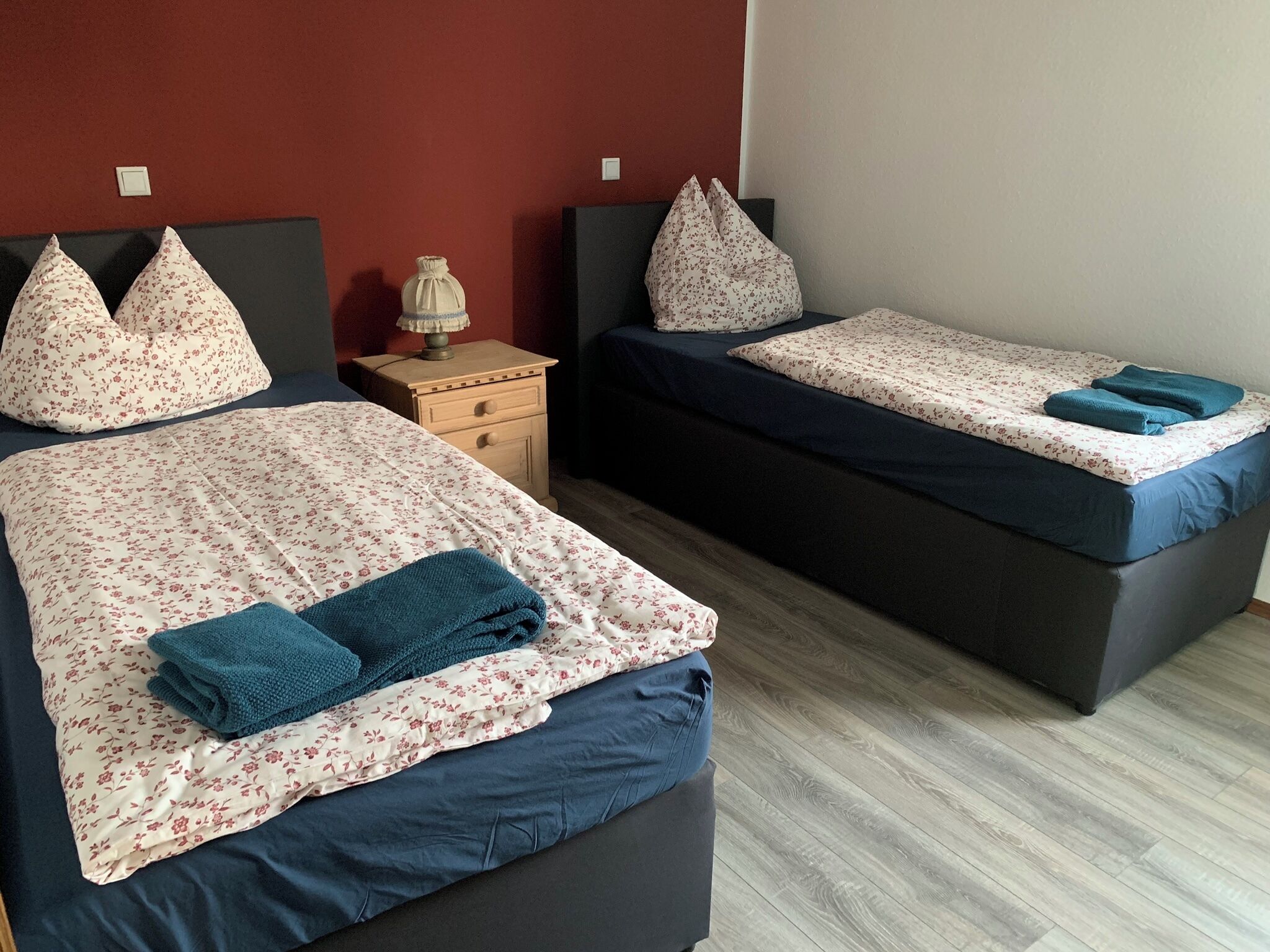 Two beds with floral bedding, a nightstand, and a lamp between them.