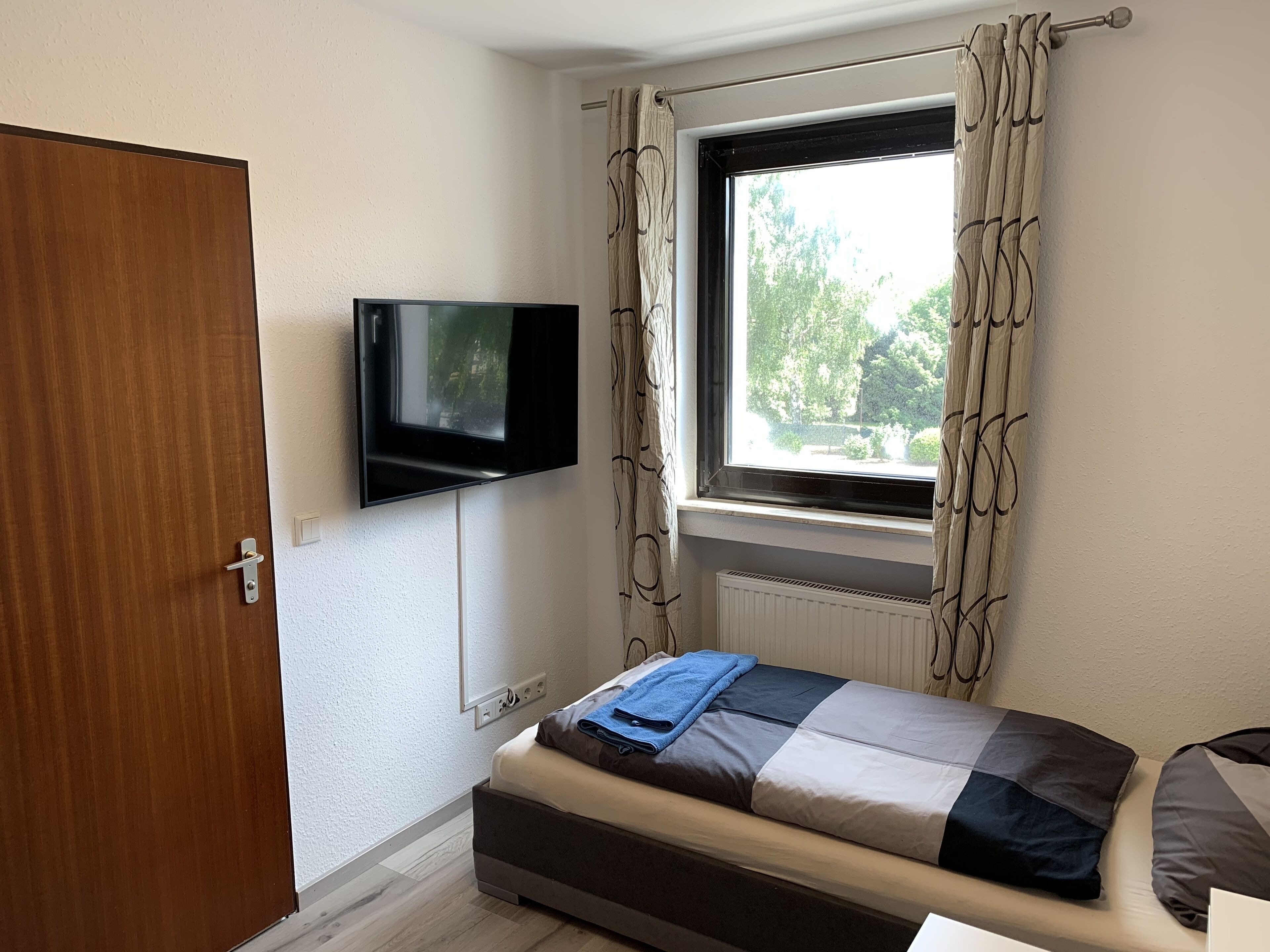 A bedroom with a bed, a TV mounted on the wall, a window with curtains, and a radiator below the window.