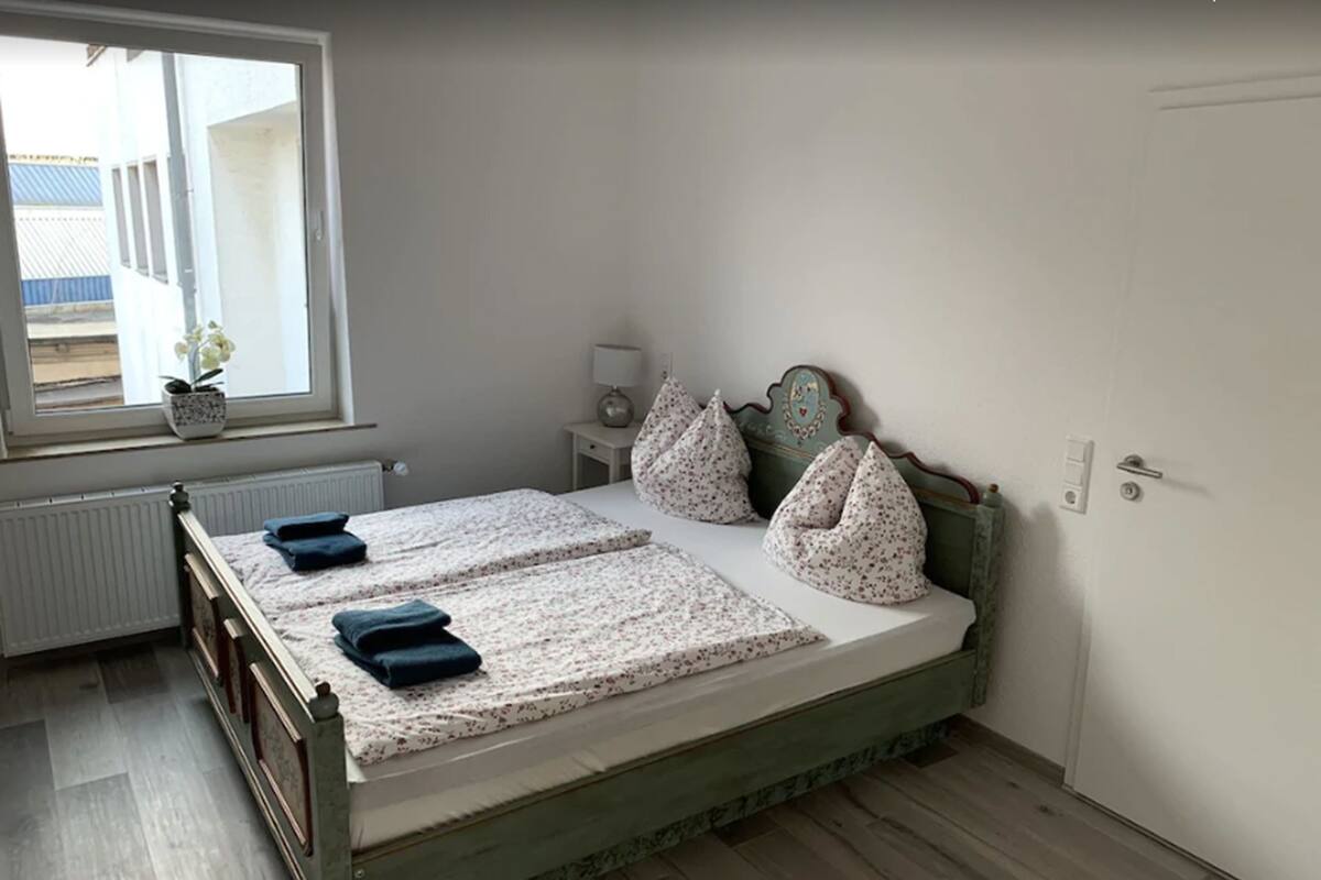 A bedroom with a bed, pillows, a bedside table with a lamp, a window with a view, and a radiator.