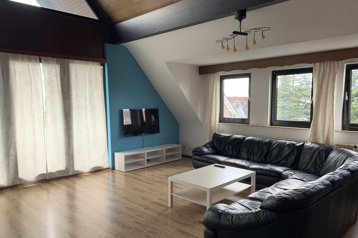 A living room with a black leather sofa, a white coffee table, a TV on the wall, and large windows.