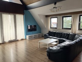 Un salón con sofá de cuero negro, una mesa de centro blanca, televisor en la pared y ventanas amplias.