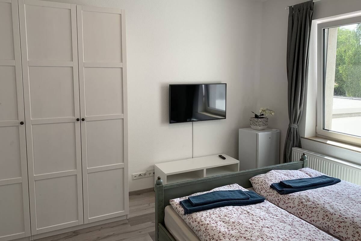 A bedroom with a bed, a TV mounted on the wall, a wardrobe, a bedside table, and a window with curtains.
