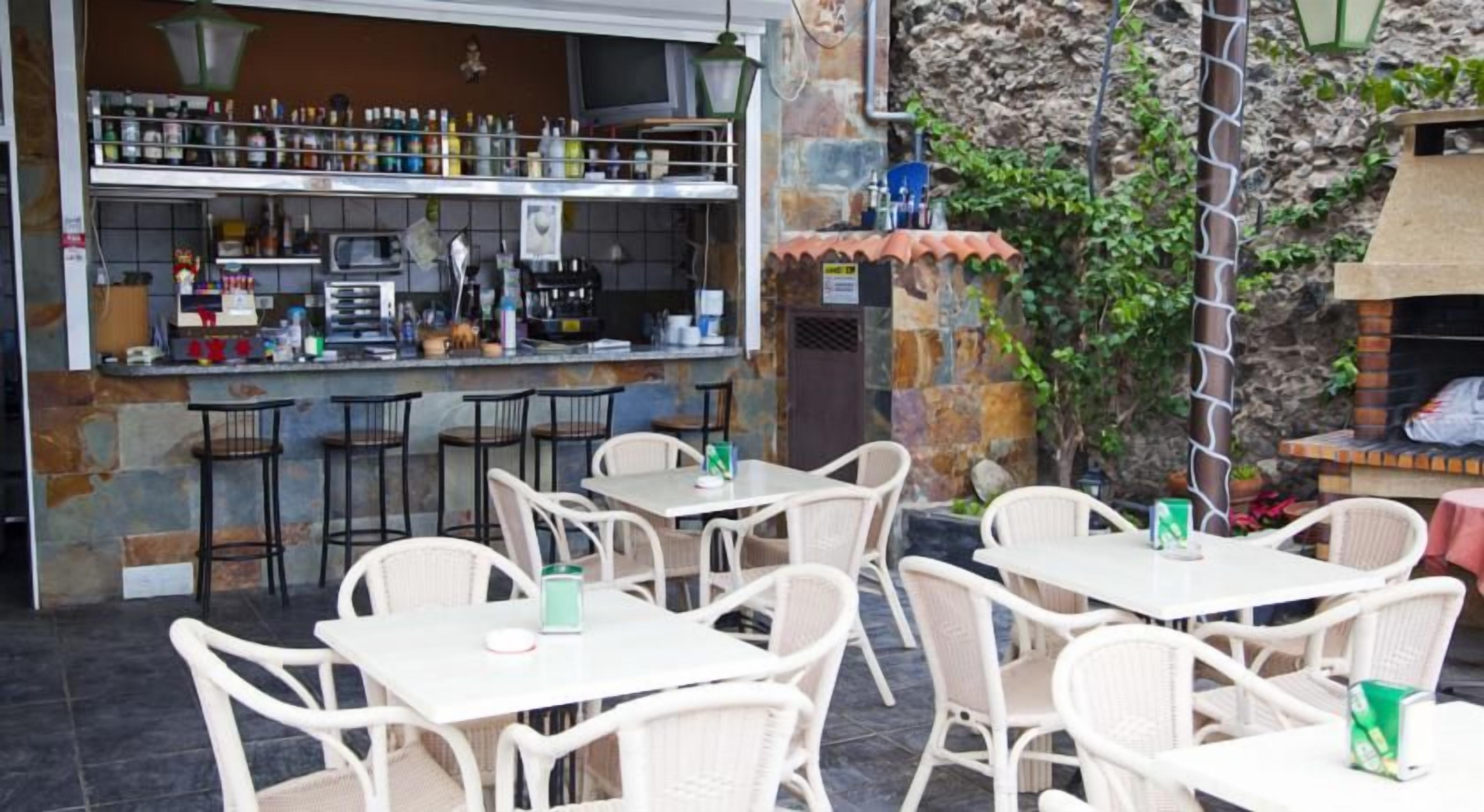 Un café al aire libre con mesas y sillas de mimbre blanco, una zona de bar con estantes llenos de botellas y un fondo de pared de piedra.