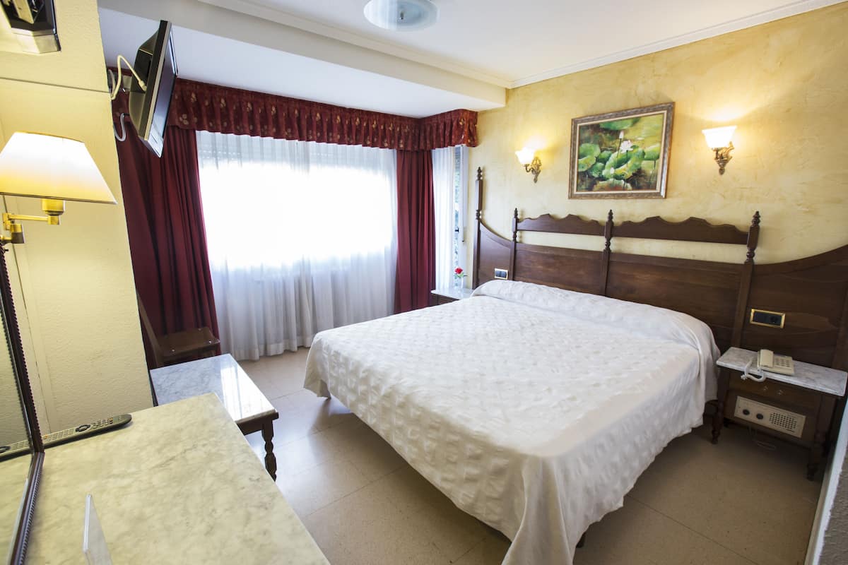 A hotel room with a large bed, a desk, a lamp, a TV mounted on the wall, and a painting above the bed.