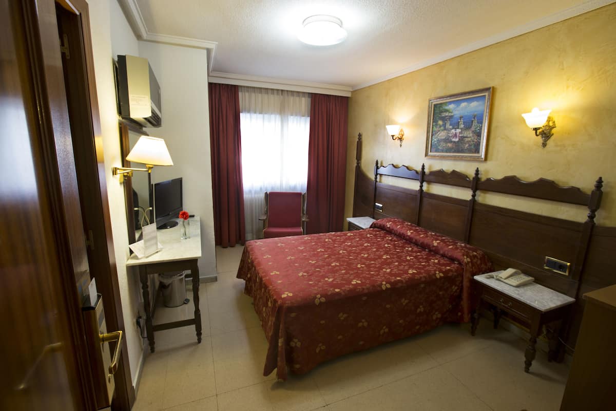 A hotel room with a bed, desk, chair, lamp, and a painting on the wall.