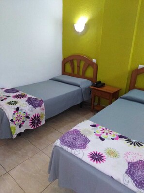 A room with two single beds, each with a wooden headboard and a grey bedspread. There is a small wooden nightstand with a lamp between the beds. The walls are painted yellow and white, and the floor is tiled. - Apartamentos Las Arenas (San Bartolomé de Tirajana)