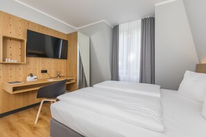 Habitación de hotel con cama, escritorio con silla, televisor montado en la pared y ventana con cortinas.