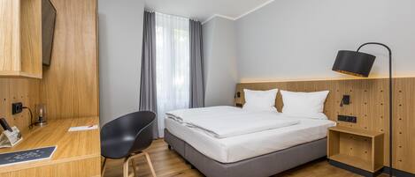 A modern hotel room with a wooden desk, a black chair, a bed with white linens, a bedside table, and a floor lamp.
