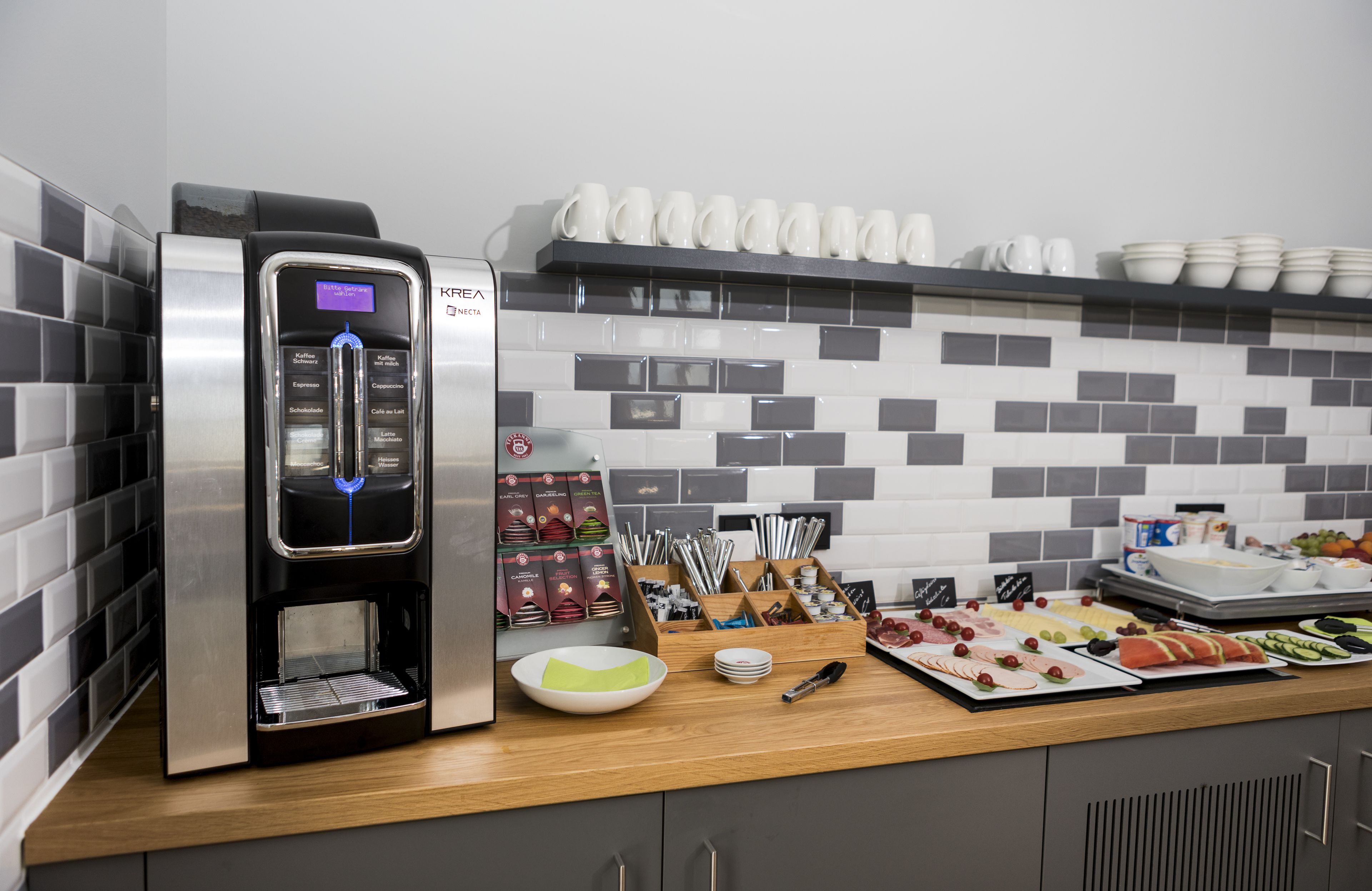 Una estación de café con una máquina de café, una variedad de tazas y un bufé con platos de comida y utensilios.