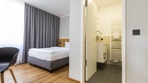 A modern hotel room with a bed, a chair, a bathroom with a toilet and sink, and a view of the outside through a window with curtains. - mk | hotel eschborn (Eschborn)