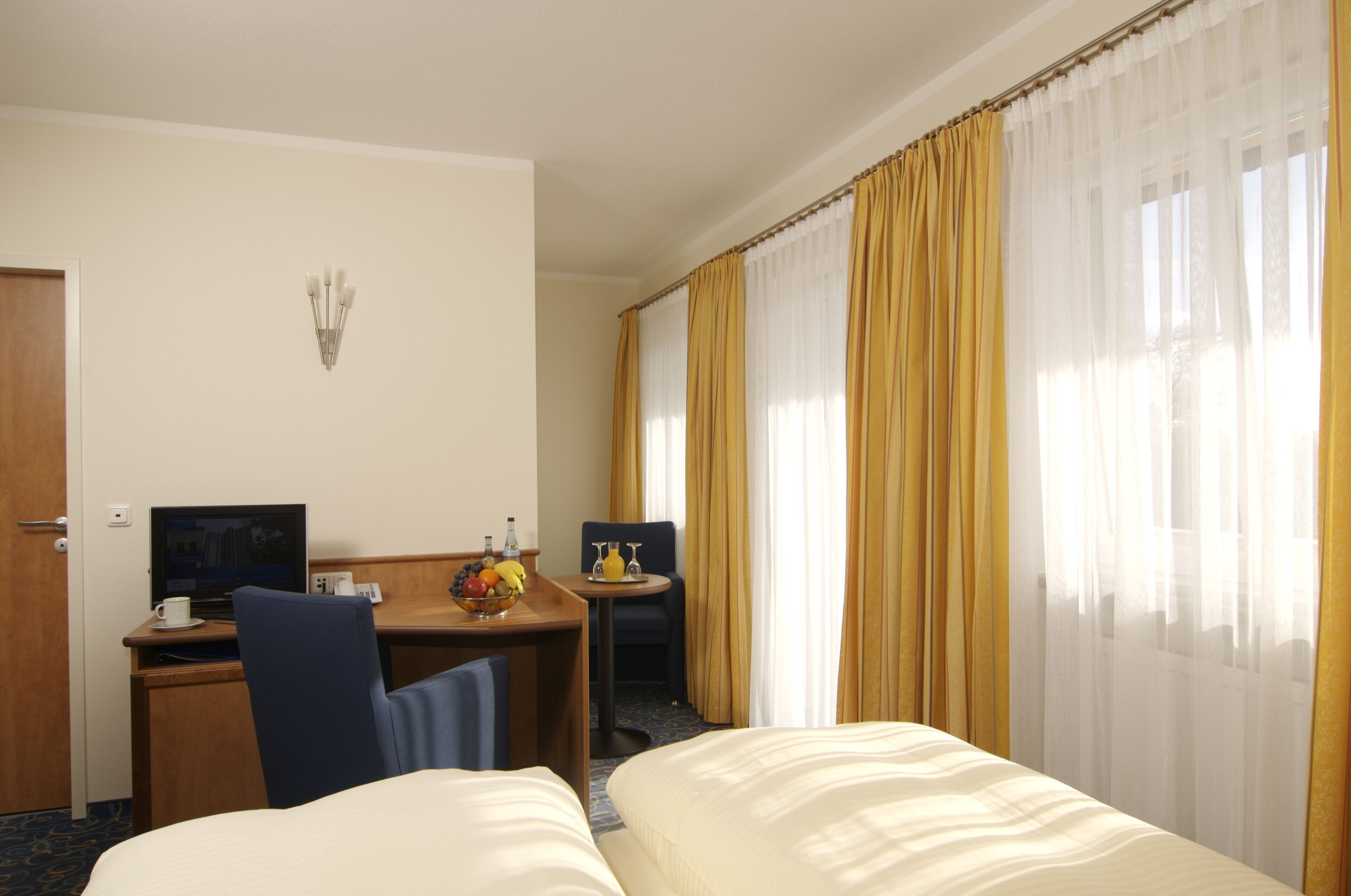 Habitación de hotel con cama, escritorio con computadora, silla, pequeña mesa con frutero y ventana con cortinas.