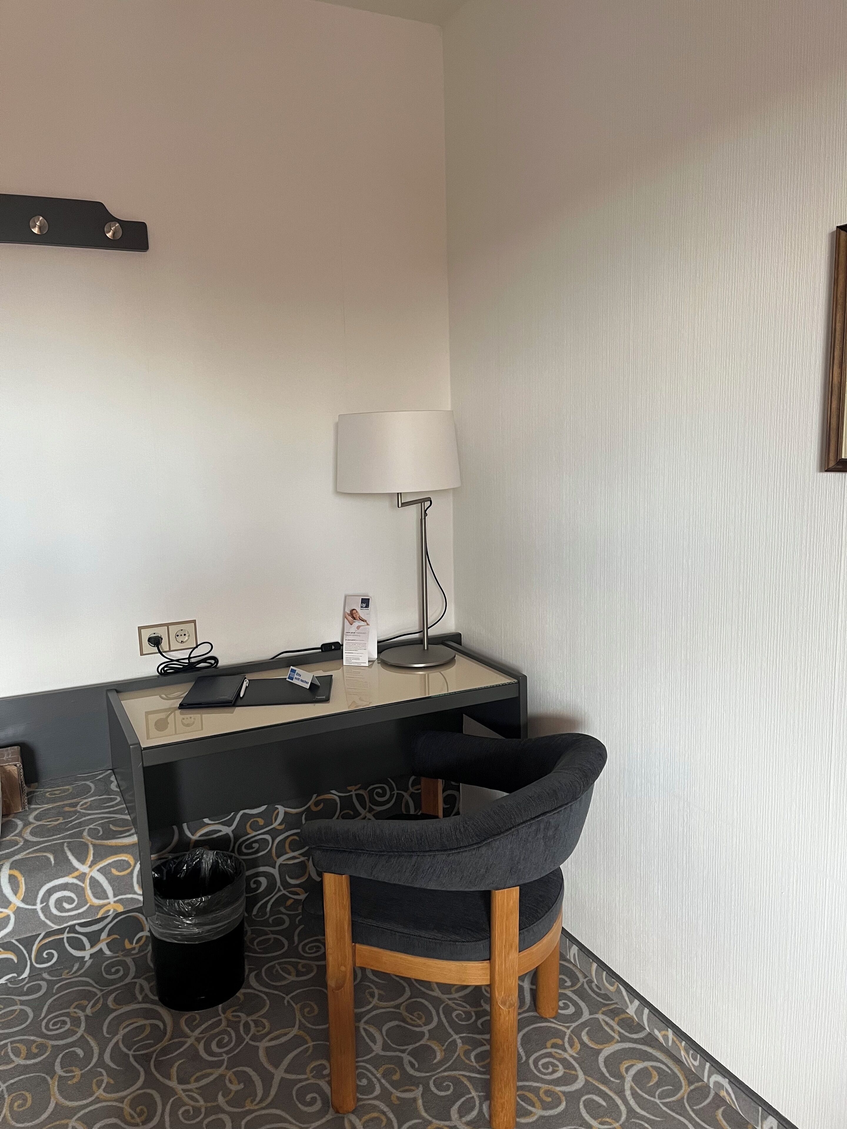 A hotel room with a desk, chair, lamp, and a trash bin.