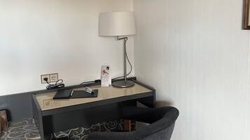 A hotel room with a desk, chair, lamp, and a trash bin.
