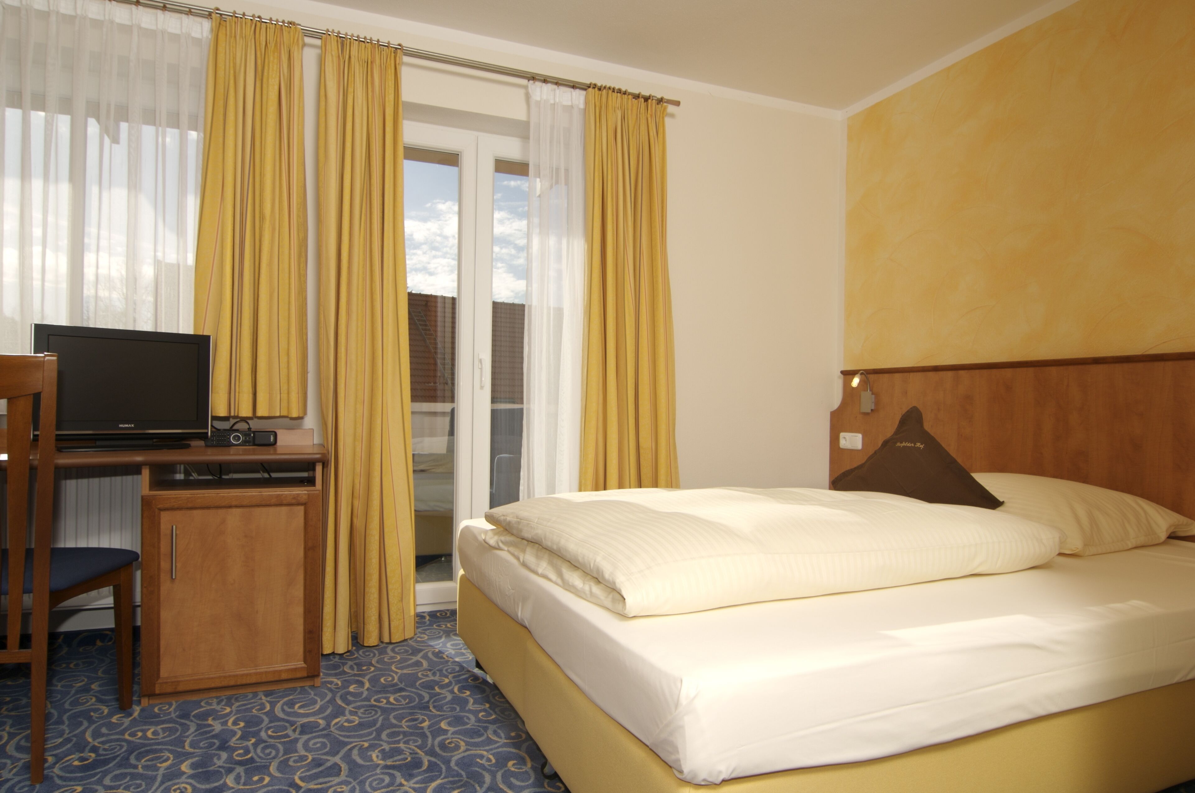 Quarto de hotel com cama, escrivaninha e televisão, e vista para o exterior através de uma janela.
