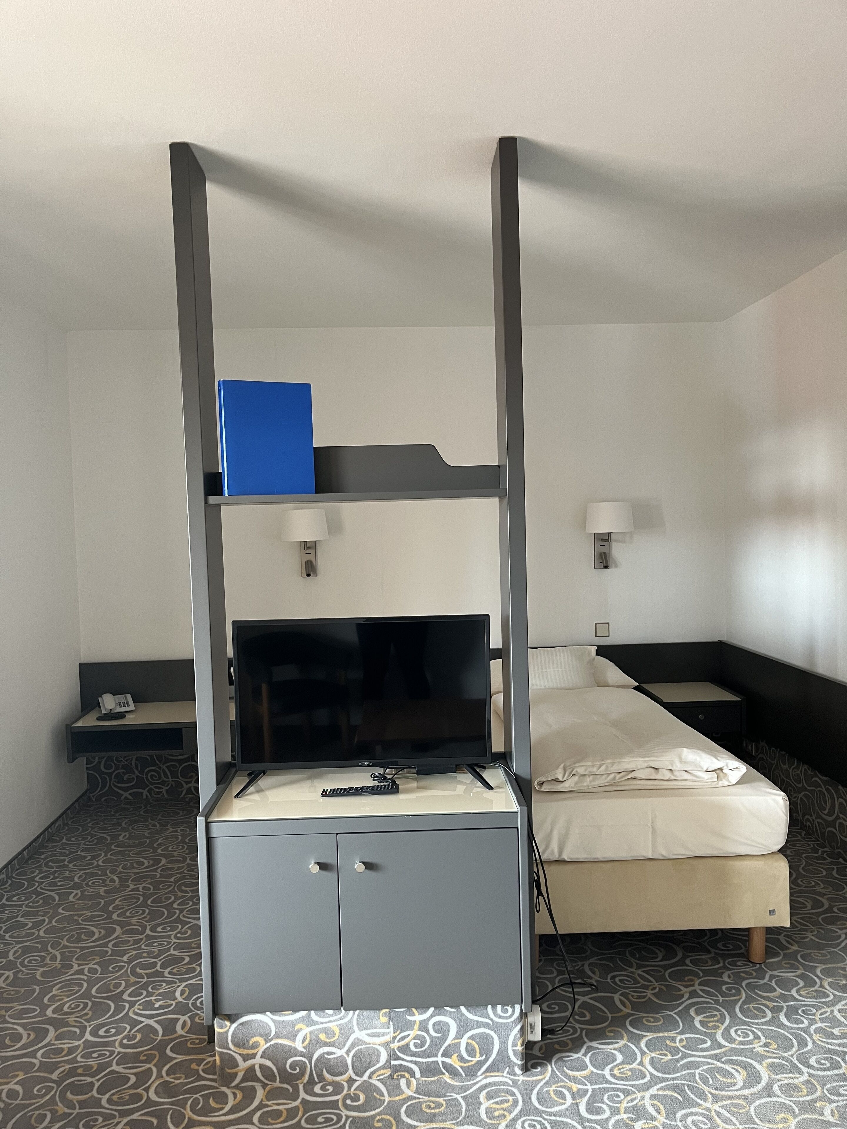 A hotel room with a bunk bed, a desk with a flat-screen TV, a lamp, and a patterned carpet.