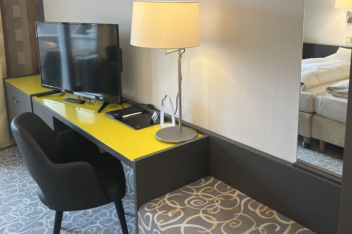 A hotel room with a yellow desk, a black chair, a television, a lamp, and a bed with white bedding.
