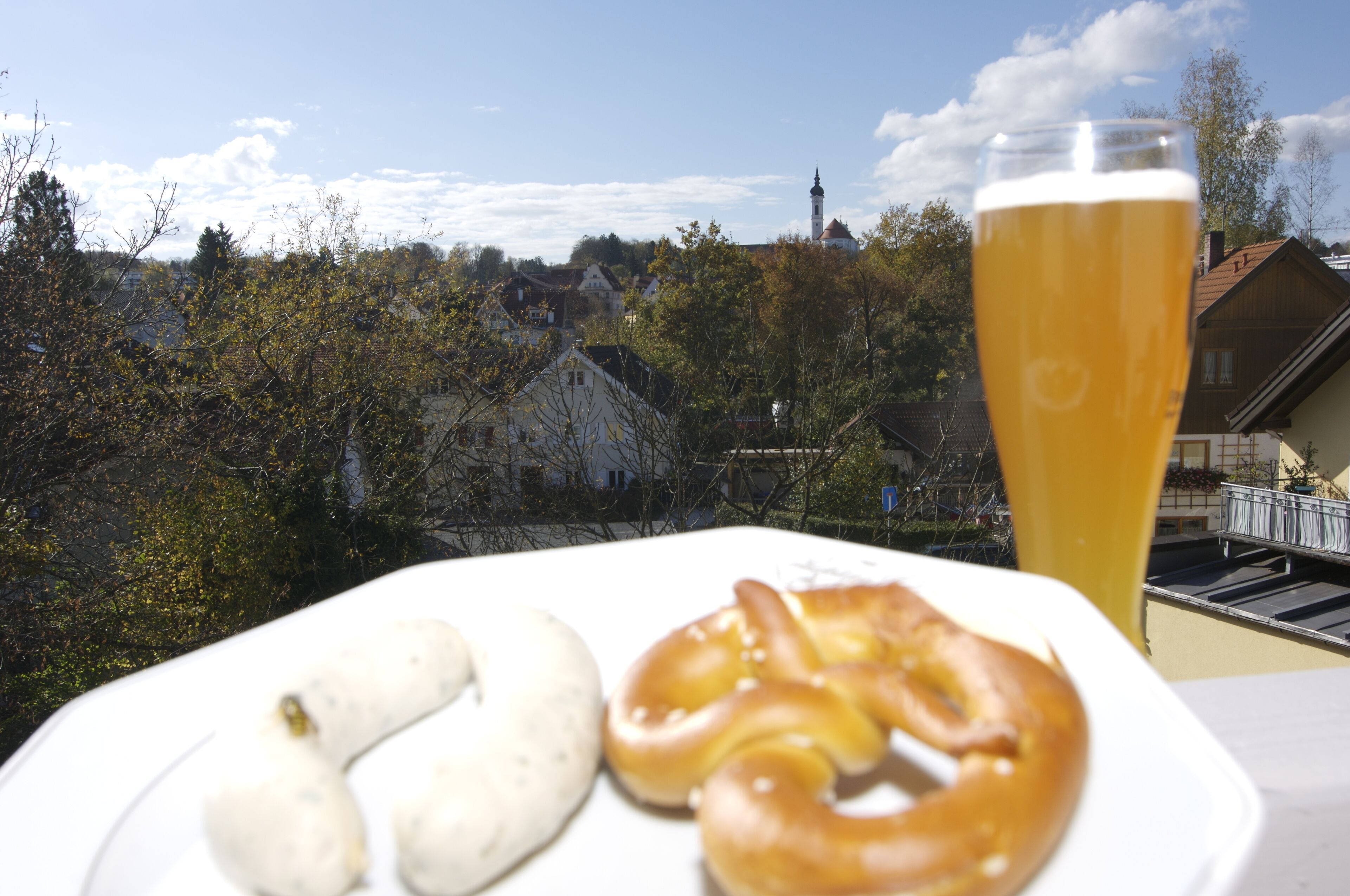 Una mesa con un plato de pretzels y salchichas, una jarra de cerveza y vista a un pueblo con una torre de iglesia al fondo.