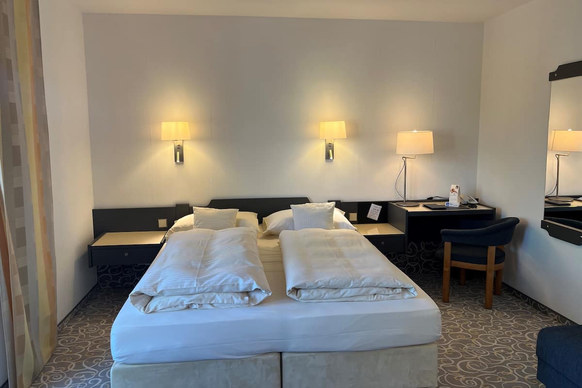 A hotel room with two beds, a desk, a chair, and two wall-mounted lamps.