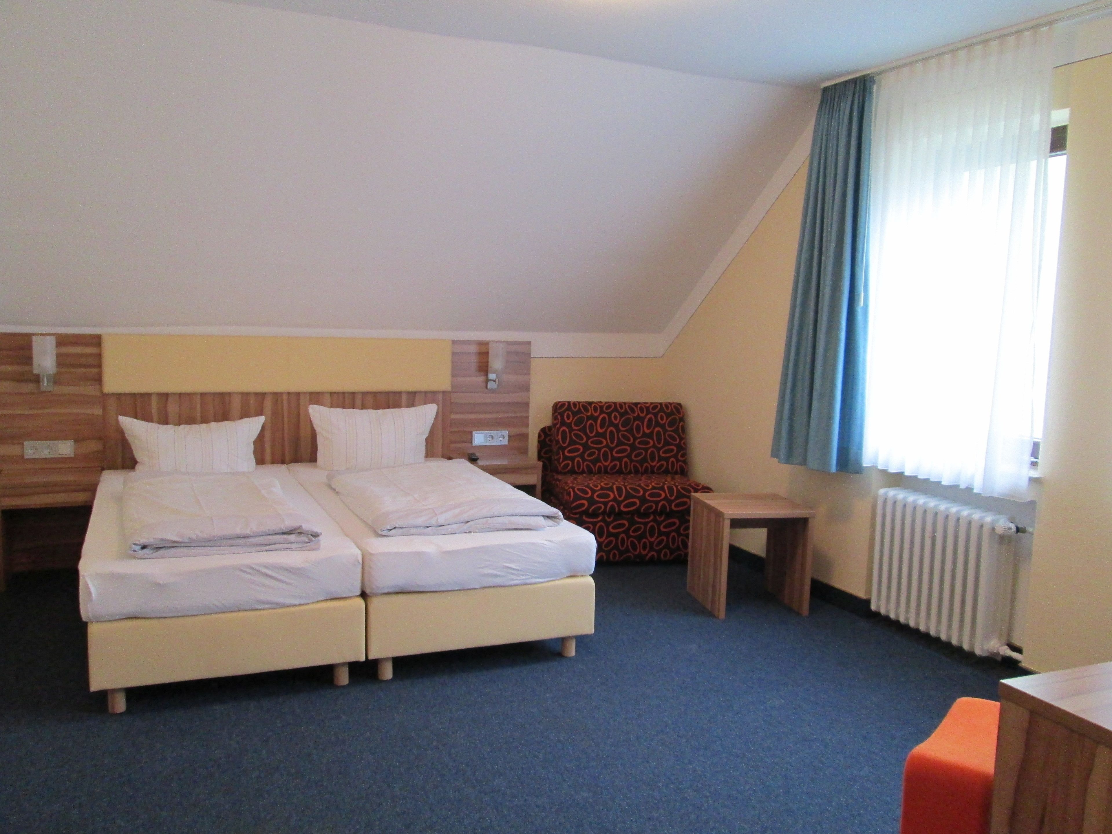 Quarto de hotel com cama de casal, uma poltrona pequena, mesa de cabeceira de madeira e uma janela com cortinas.
