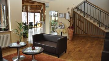 A hotel lobby with a leather sofa, two small tables, a large potted plant, a staircase, and a reception area with a glass display case.