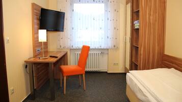 Quarto de hotel com escrivaninha de madeira, uma cadeira, uma cama, televisão na parede e janela com cortinas.
